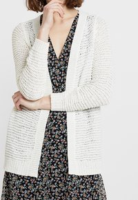 Žena v bílé otevřené pletené cardiganě přes černé květinové šaty s výstřihem do V, se založenýma rukama a jednou rukou blízko brady.