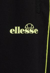 Svarta träningbyxor med en gul "ellesse"-logotyp och en rand längs sidan, tillverkade av ett slätt, strukturerat tyg.
