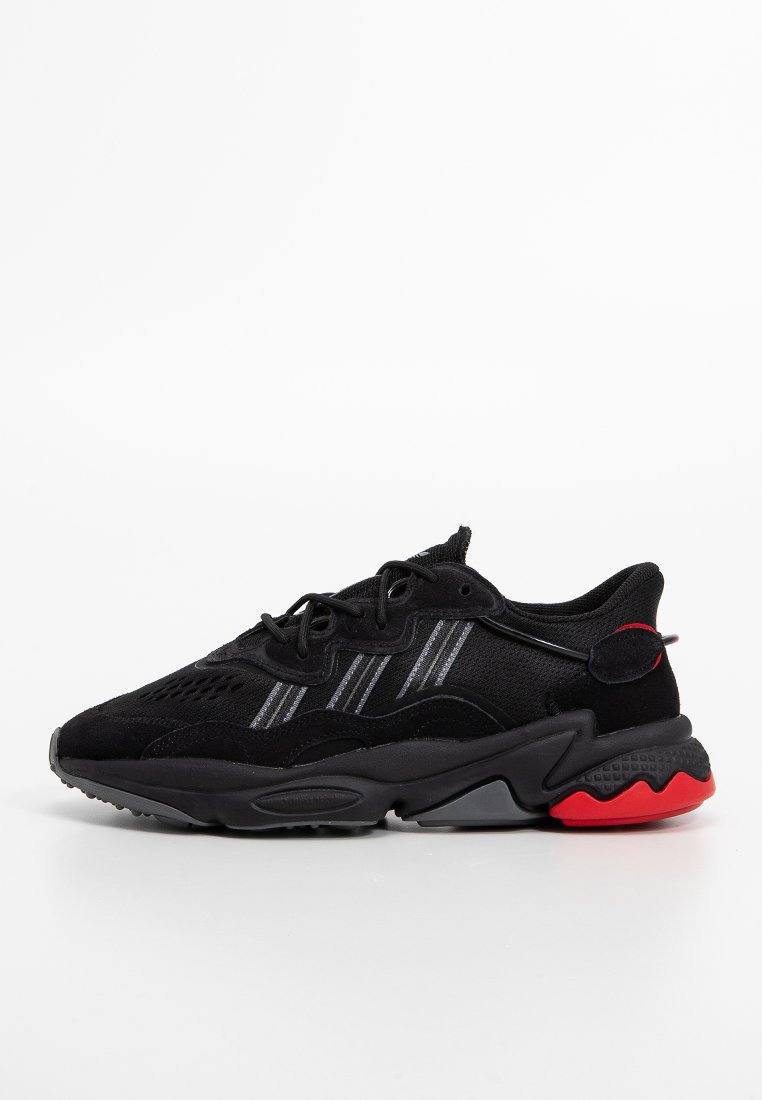 Schwarze Sportschuhe mit Obermaterial aus Mesh und Wildleder, grau gestreifte Akzente, strukturiertes schwarzes Zwischensohlenmaterial und rote Außensohle. Abgerundete Zehenform und Schnürverschluss.