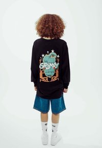 Svart långärmad tröja med grafisk design i gröna inslag och ordet "GRIMY" på ryggen, tillsammans med denimshorts och vita strumpor.