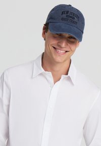 Granatowa czapka z wyszywanym napisem "NEW JERSEY EAST COAST" na czarno, miękka tkanina, wygiębrim, regulowany pasek z tyłu. Noszona z białą koszulą.