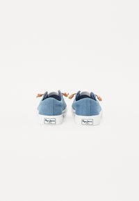Ténis de lona slip-on em azul claro com solas de borracha brancas, apresentando atacadores castanhos e uma etiqueta de marca no calcanhar.