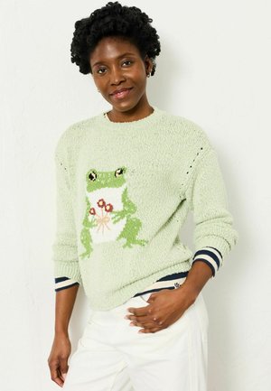 Smiling woman with short curly hair wearing a light green sweater featuring a frog holding red flowers, paired with white pants.