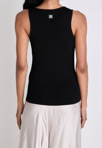 Donna che indossa un top nero a coste senza maniche con un piccolo logo in metallo quadrato sulla parte superiore della schiena, abbinato a pantaloni in satin di colore chiaro.