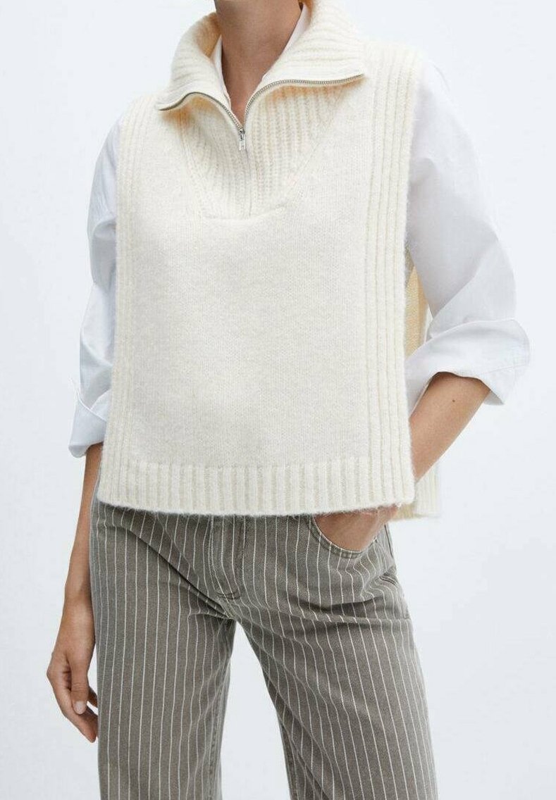 Elfenbeinfarbener, gestrickter ärmelloser Pullover mit hohem Kragen, mit V-Ausschnitt und geripptem Muster. Kombiniert mit gestreiften grauen Hosen.