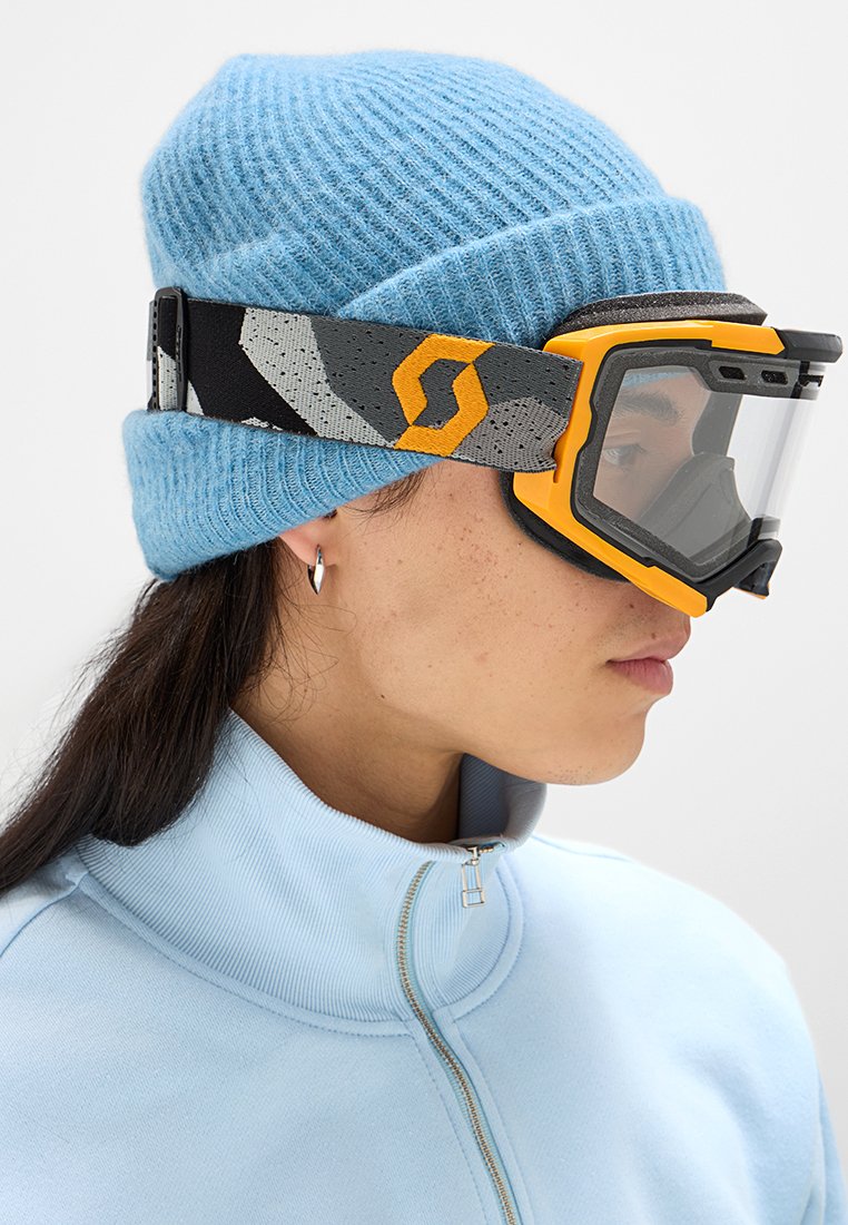 Gorro de lana azul, tira de gafas de esquí negra y gris con logo naranja, y gafas de esquí transparentes; llevadas con una chaqueta de cremallera azul claro.