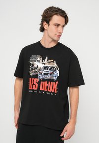 Čierne bavlnené tričko s grafickou potlačou pretekárskych áut a panorámy mesta, s červeným nápisom „LES DEUX“ v tučnom písme. Krátke rukávy a voľný strih.