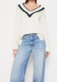 Kvinna iklädd en vit ribbad tröja med svarta V-formade ränder, matchad med höga, ljusblå vida jeans mot en neutral bakgrund.