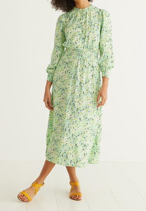 Vrouw draagt een lichtgroene midi-jurk met bloemenprint, lange mouwen, een geplooide taille en gele sandalen, staand op een witte vloer.
