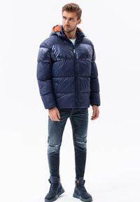 Marineblauwe pufferjack met oranje voering in de capuchon, ritssluiting aan de voorkant, geborduurd ontwerp en geribbelde manchetten, gecombineerd met distressed jeans en marineblauwe hoge sneakers.