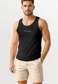 Schwarzes ärmelloses Tanktop mit rundem Ausschnitt, mit weißem Schriftzug "Paradise" in der Mitte. Kombiniert mit hellbeigen Shorts.