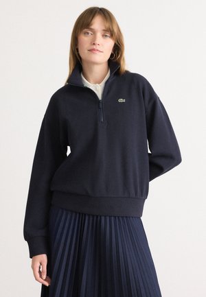 Navy zip-up sweatshirt in soft fabric with ribbed hem and cuffs. Features a green logo on the chest. Worn over a pleated navy skirt.