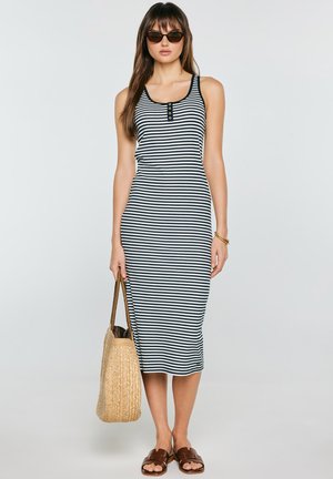 Kobieta w czarno-białej pasiastiej sukience midi bez rękawów, okulary przeciwsłoneczne, brązowe sandały, złota bransoletka i trzyma plecioną słomianą torbę.