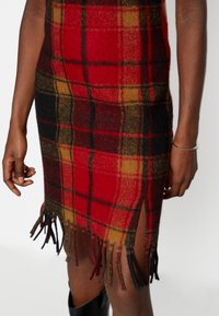 Jupe en laine à carreaux rouge et noir avec un ourlet à franges, dotée d'une fente sur le côté. Le tissu texturé mélange des couleurs rouge, jaune et noir.