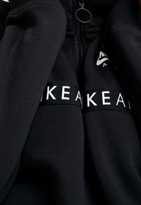 Svart hoodie med dragkedja, försedd med en logo på bröstet och en kontrasterande vit textrem. Mjukt tyg och avslappnad passform.