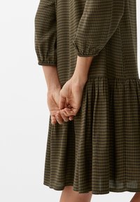 Kleid aus braunem und grünem Gingham mit dreiviertellangen Puffärmeln, geraffter Taille und schwingendem Rock; der Stoff ist leicht und hat eine glatte Textur.