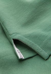 Gros plan d'une manche en tissu vert texturé avec une petite étiquette blanche partiellement visible à l'intérieur du poignet.