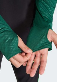 Hosszú ujjú ing sötétzöld texturált anyagból, hüvelykujjszegélyekkel, fekete anyaggal alatta. Simogató, testhezálló kialakítás.