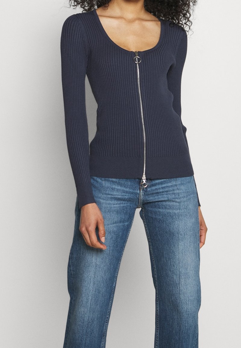 Mörkblå ribbad långärmad cardigan med scoop-halsringning och central dragkedja med ett cirkulärt drag, matchad med ljusblå vida jeans.
