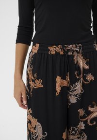 Persoon in een zwarte top met driekwartmouwen en zwarte broek met bruine en beige paisley-patronen, vastgebonden in de taille met een koord.