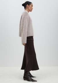 Woman with dark hair in a bun wearing a fuzzy light gray sweater, long dark brown skirt, and black pointed boots standing in profile.