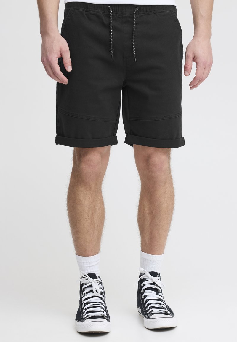 Shorts en coton noirs avec une taille à cordon de serrage, des ourlets roulés, des coutures latérales et une coupe décontractée, portés avec des baskets montantes noires et des chaussettes blanches.