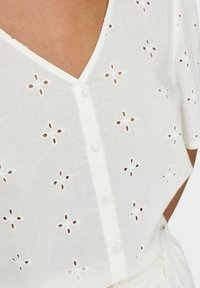 Hvid bomuldsbluse med V-udskæring, korte ærmer og knaplukning, dekoreret med broderede hulmønstre af blomster.