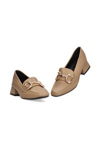 Mocasines de imitación piel en color beige con un bajo tacón cuadrado, que presentan un acento de cadena dorada en el empeine y detalles de costura visibles.