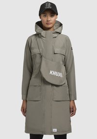 Parka lung, de culoare gri deschis, cu guler înalt, multiple buzunare frontale și închidere cu fermoar. Include o geantă crossbody de culoare khaki cu "KHUJO" imprimat în alb.