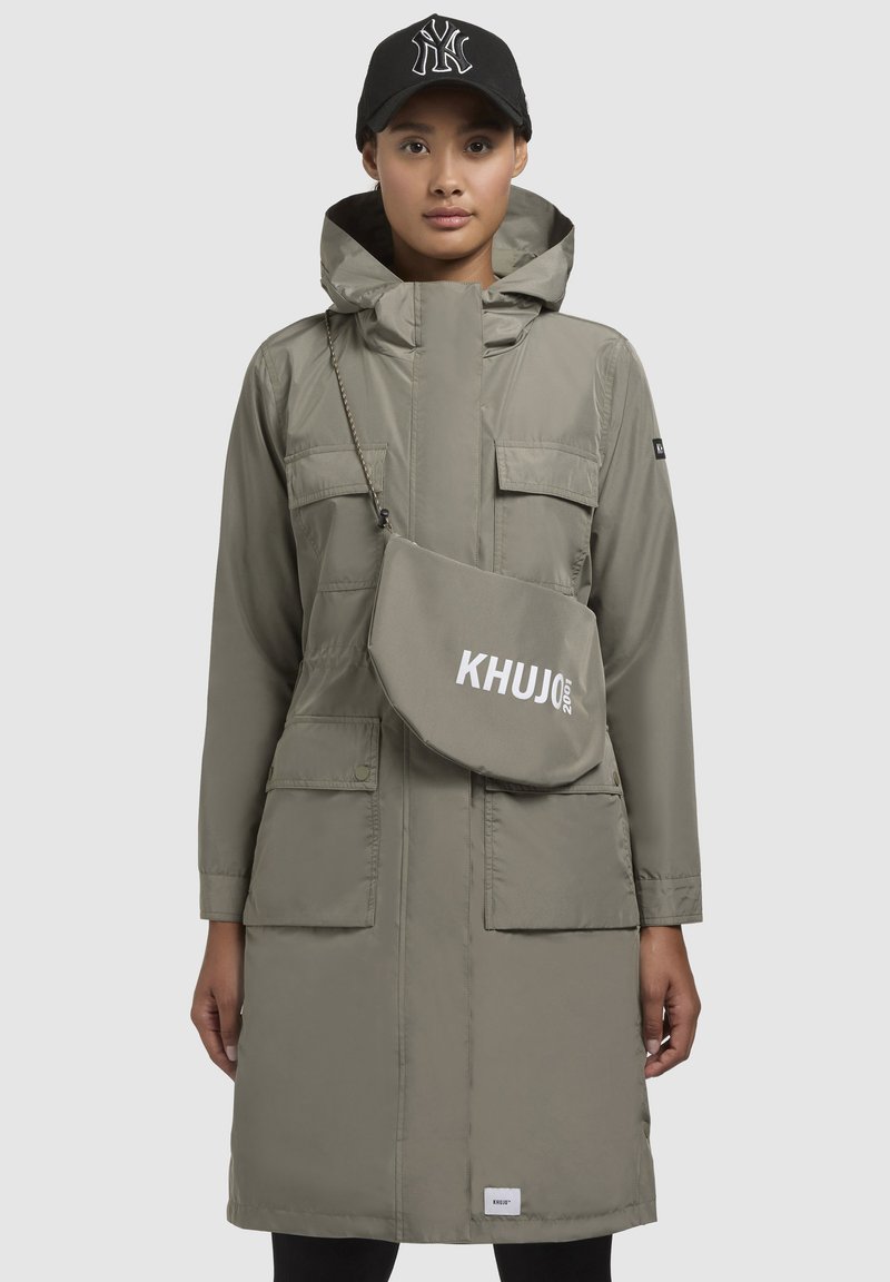 Pikk, heledat hall parka koos kõrge kraega, mitme esikaanega tasku ja tõmbluku sulgemisega. Kaasa kuulub khakivärvi rippkotid, millel on valge "KHUJO" trükk.