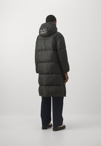 Lang sort puffer frakke med hætte, præget af et quiltet design og en blank tekstur. Båret over mørke bukser og solide sko.