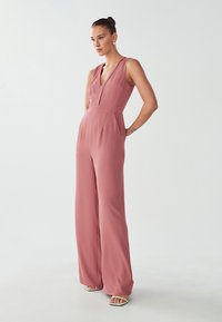 Blush rozenkleurige jumpsuit met een diepe V-hals, mouwloos ontwerp en wijde broekspijpen. Gemaakt van een gladde, lichte stof.