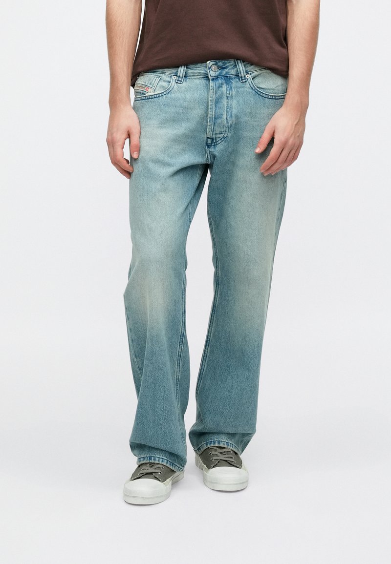 Ανοιχτό μπλε τζιν, άνετη εφαρμογή, ξεθωριασμένο μπροστινό μέρος, σχεδιασμός με πέντε τσέπες, μεταλλικά εξαρτήματα και ραφές λεπτομέρειες. Φορεμένο με γκρι αθλητικά παπούτσια.