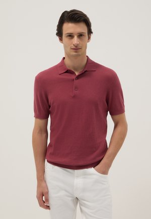 Hombre con una camisa polo de manga corta color burdeos y pantalones blancos, de pie contra un fondo claro y liso.