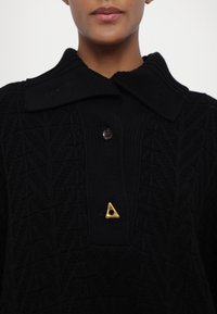 Zwarte gebreide sweater met een grote kraag, voorzien van een gestructureerd patroon en een gouden driehoekige speld als accent. Twee bruine knopen aan de voorkant.