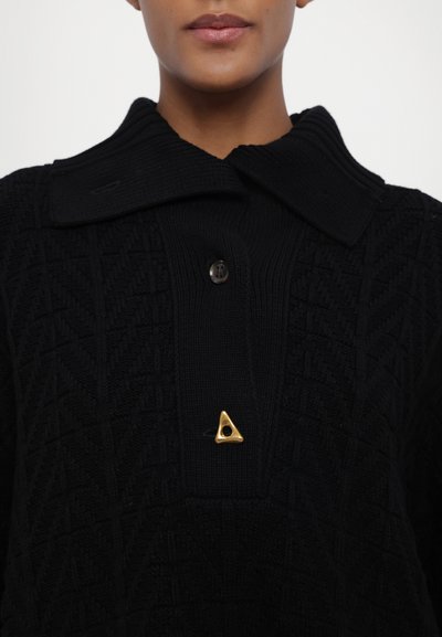 Black knitted sweater with a large collar, featuring a textured pattern and a gold triangular pin accent. Two brown buttons on the front.