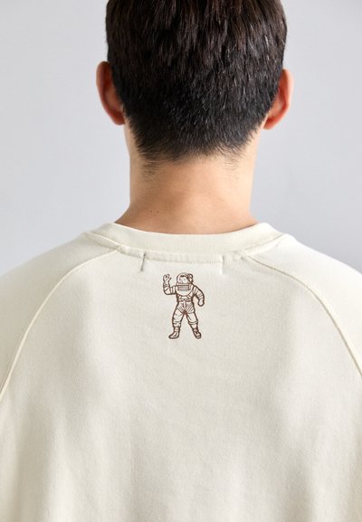 Back of person’s head with short dark hair wearing a cream sweater featuring a small astronaut graphic near the neckline.