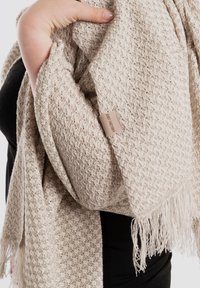 Beige geweven sjaal met een gestructureerd patroon, franjes aan de randen en een klein merklabel. Zachter materiaal dat over een schouder hangt.
