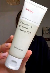 Mano sosteniendo un tubo de gel exfoliante con enzima galactomy de la marca Manyo, etiquetado como "Exfoliante Suave", sobre un fondo interior desenfocado.