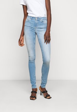 Kvinde iført lyseblå skinny jeans, hvid kortærmet skjorte og sorte høje hæle med flettede stropper, stående indendørs.