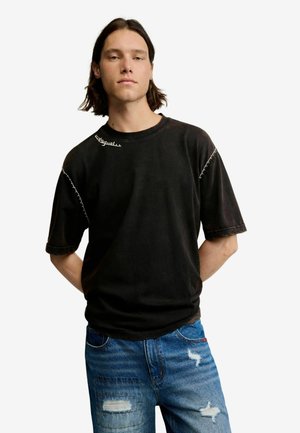 Junger Mann mit schulterlangem Haar, der ein schwarzes übergroßes T-Shirt und blaue, abgenutzte Jeans trägt und mit den Händen hinter dem Rücken steht.