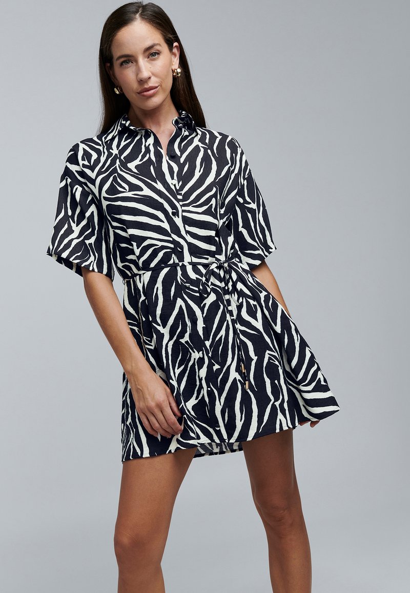 Schwarz-weißes Kleid mit Zebramuster, kurzen Ärmeln, Knopfleiste vorne, tailliert und locker geschnitten. Texturiertes Gewebe mit leichtem Glanz.