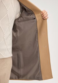 Persoon in een beige gebreide trui en broek houdt een tan jas open, waardoor de soepele donkergrijze binnenvoering met een ritszak zichtbaar wordt.