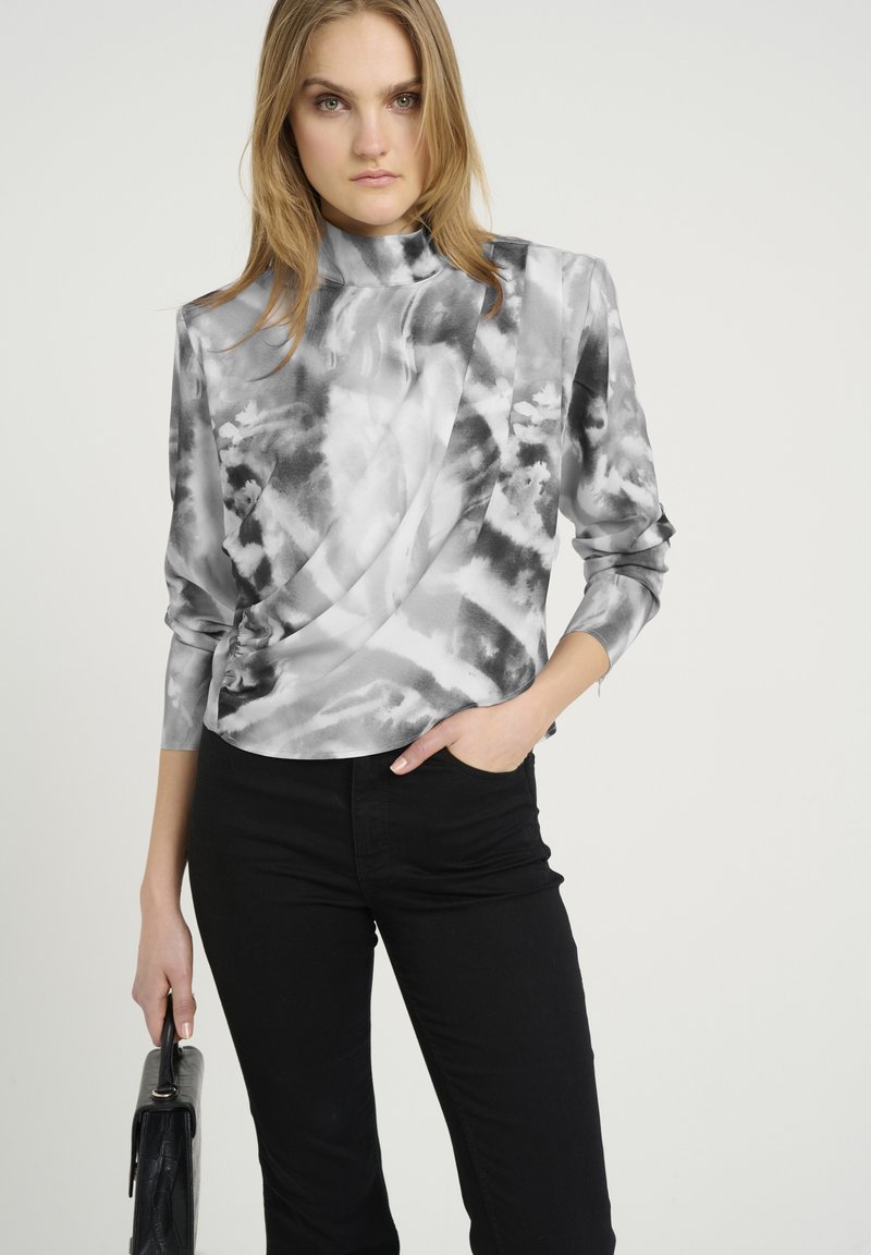 Top lungo a maniche lunghe grigio e nero con collarino alto, design drappeggiato e texture morbida, abbinato a pantaloni neri e una borsa nera.