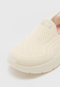 Vaalean beige liukuva tennari, jossa on hengittävä verkkokuori, pehmeä välipohja ja hillitty tekstuuri. Sivulla on brändätty merkki.