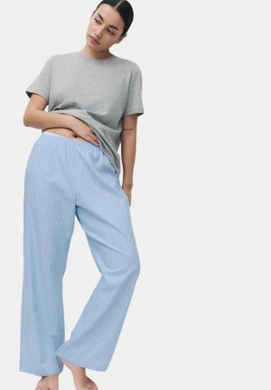 Donna che indossa una t-shirt grigia e pantaloni larghi a righe azzurro chiaro, in piedi a piedi nudi con una mano che solleva leggermente la maglietta.