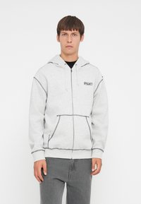 Grå zip-up hettegenser med lomme foran, kontrasterende sømmer og et logo-merke. Har hette og elastiske mansjetter. Brukt med grå jeans.