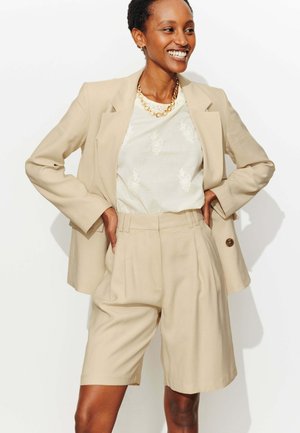 Femme souriante, portant un blazer beige et un short taille haute assorti sur un haut blanc à motif, accessoirisée d'un collier à chaîne en or.