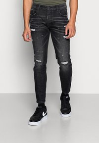 Jeans vaqueros negros con roturas desgastadas, diseño de corte ajustado y detalles plateados; combinados con zapatillas negras con un logo blanco.