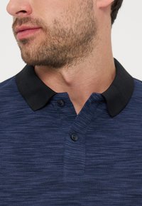 Άνδρας με ελαφριά γενειάδα που φορά μπλε πικέ πόλο με μαύρο γιακά, τα κουμπιά είναι κουμπωμένα κοντά στον λαιμό, κοιτάει προς τα αριστερά.